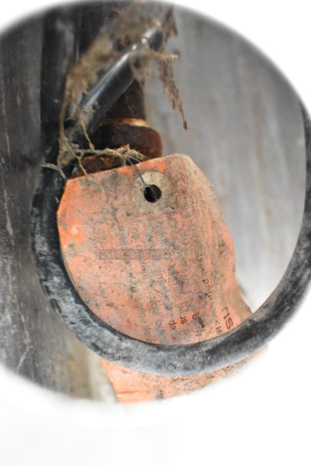 A close-up of a worn, orange tag with text, attached to a black wire and metal surface, with dirt and wear visible.