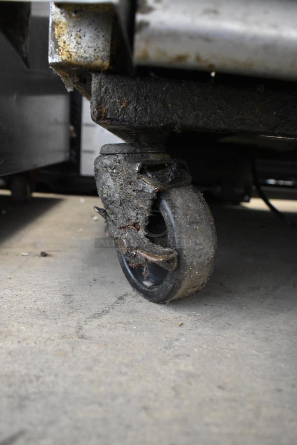 Malformed, rusty caster of a True T-23 stainless steel commercial cooler. Signs of heavy use, impacting mobility.