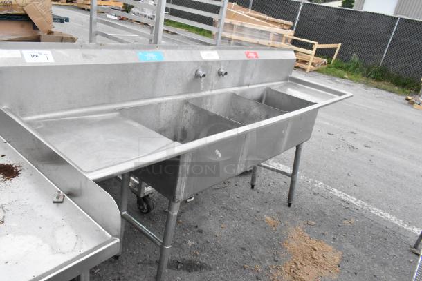 Stainless steel 3 bay sink featuring dual drain boards, used condition. Ideal for commercial kitchens. Visible wall-mounted taps.