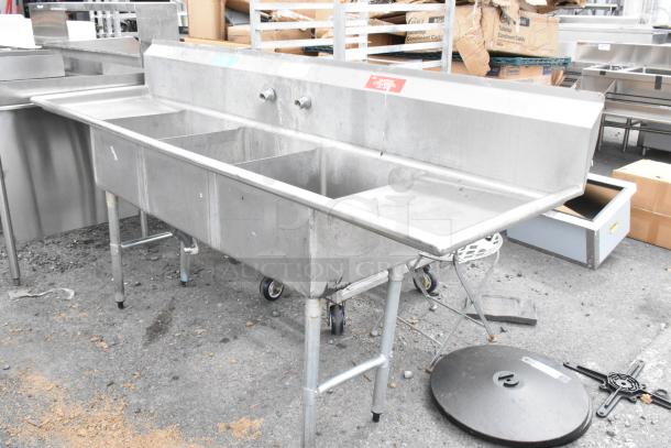 Stainless steel 3 bay sink with dual drain boards, visible signs of wear, faucets and plumbing attached, commercial use.