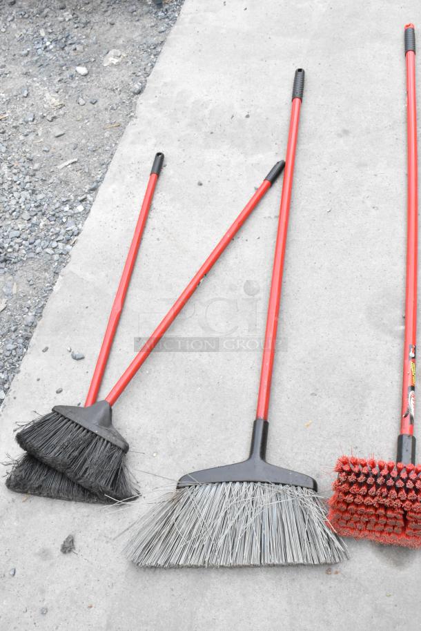 Set of three brooms with red handles, featuring various bristle styles, displayed on concrete surface.