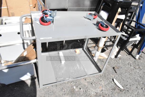 Brand new scratch and dent gray metal 2-tier cart with commercial casters. Features flat top, lower shelf, and sturdy frame.