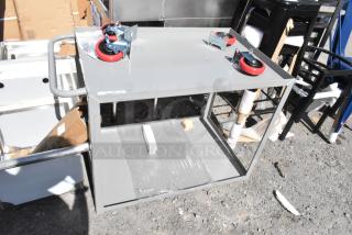 Brand new scratch and dent gray metal 2-tier cart with commercial casters. Features flat top, lower shelf, and sturdy frame.
