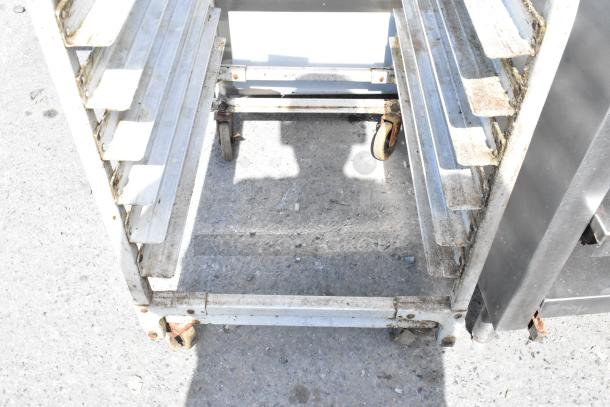Metal pan transport rack on commercial casters, shows signs of wear, rust visible on wheels. Suitable for kitchen use.
