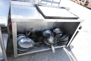Stainless steel table with under shelf, holding assorted skillets. Slight wear on surface, ideal for kitchen use.