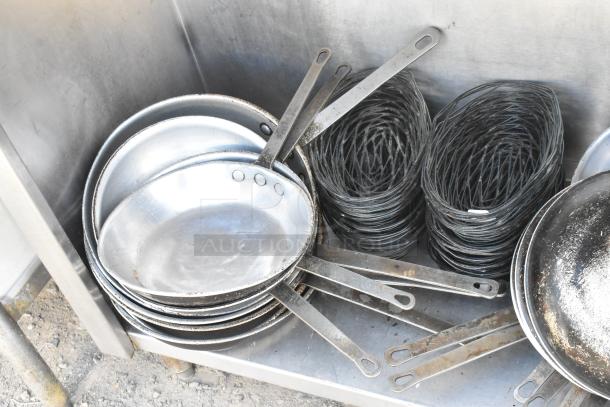 Stainless steel table with under-shelf containing a stack of skillets and metal coils. Used kitchen equipment in fair condition.