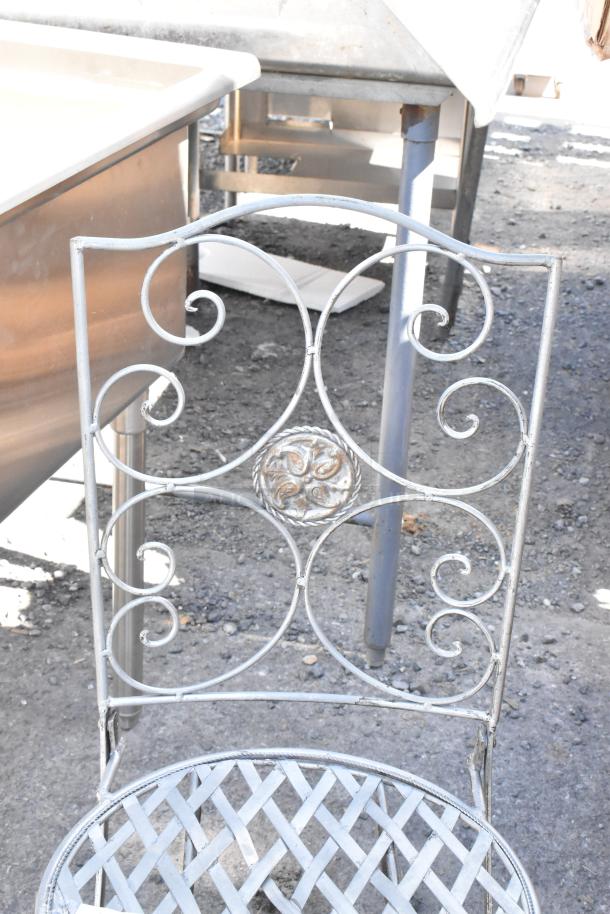 Black metal wire patio chair with ornate scroll design, circular medallion on backrest, lattice seat, slight wear.