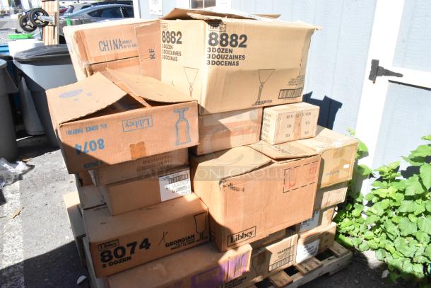 Boxes of brand new scratch and dent dishware, including Libbey glasses, stacked on pallets outdoors. Visible box text indicates contents and quantities.