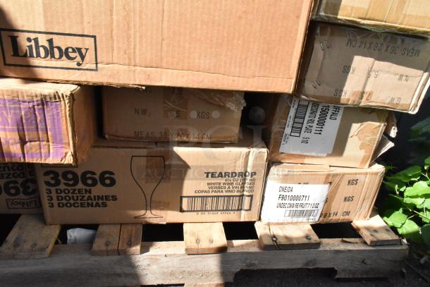 Boxes labeled with brands like Libbey and Oneida on a pallet, containing new scratch and dent dishware, including wine glasses.