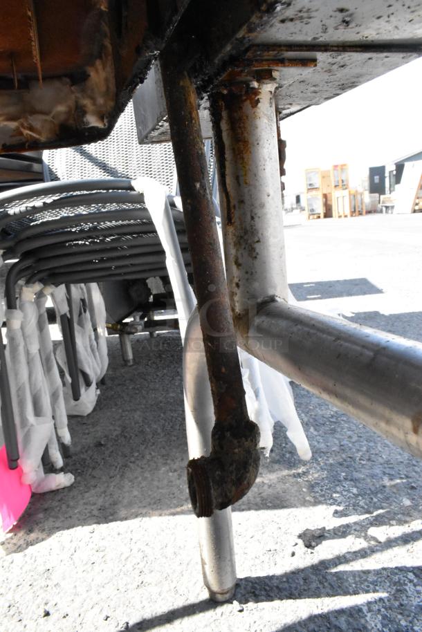 Stainless steel commercial manual tilt braising pan underside, showing legs and connections. Signs of rust and wear visible.