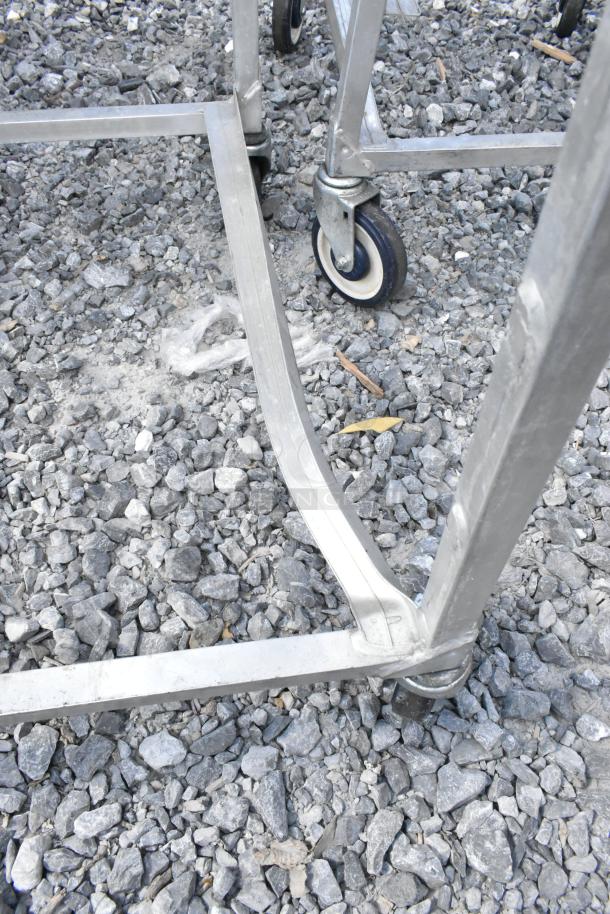 Two metal commercial pan transport racks on casters, shown on gravel surface. Durable construction, ideal for kitchen use.