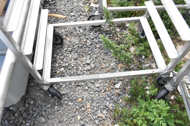 Two metal commercial pan transport racks on commercial casters, sturdy design, suitable for kitchen use, shown on gravel.