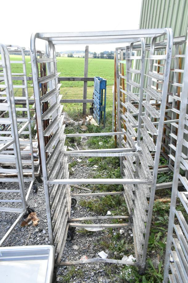 Two metal commercial pan transport racks on commercial casters, outdoor setting, showing visible wear and sturdy design.