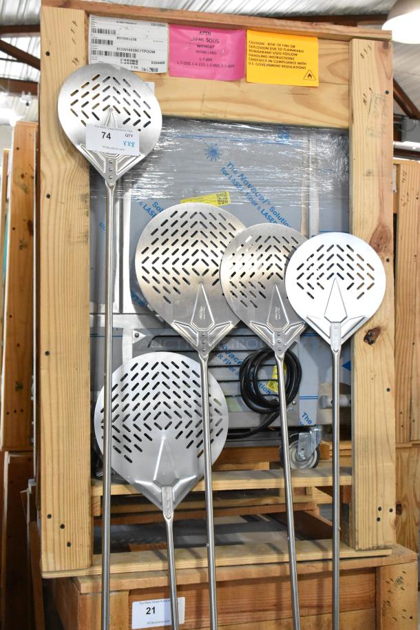 Nine brand new perforated pizza peels in various sizes, stainless steel finish, labeled as scratch and dent, on wooden pallet.