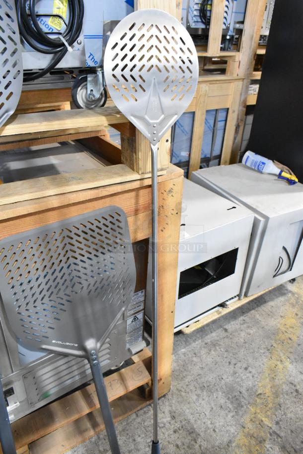 Nine brand new scratch and dent, various sized pizza peels with perforated blades, metal handles, in a warehouse setting.