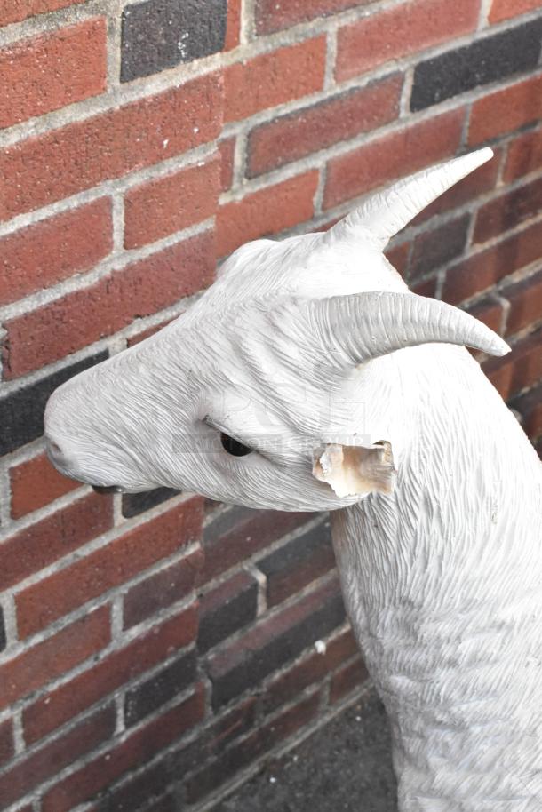 "White goat statue, realistic texture, horn detail, slight damage on ear, against brick wall. Auction includes cow statue."