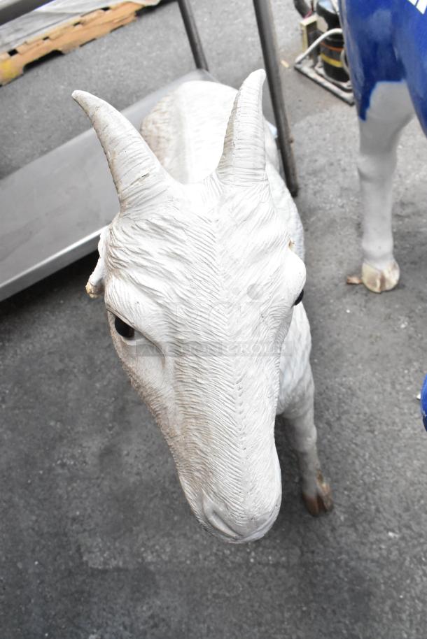 White goat statue with detailed texture, standing outdoors. Visible wear on horns and nose; background shows partial cow statue.