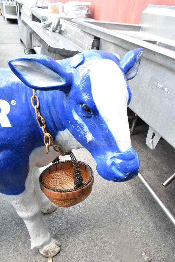 Blue and white cow statue with a wicker basket, chain around neck, in a metal-industrial setting.