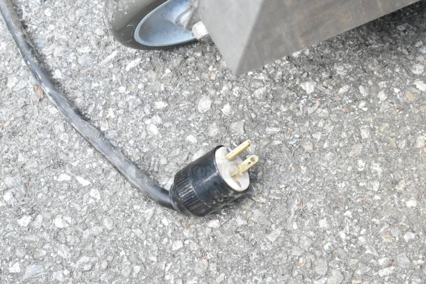 Close-up of a black electric plug with a worn cable on an asphalt surface near a metal table leg on a caster wheel.