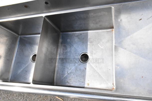 Stainless steel 3-bay sink with dual drain boards, lacking legs; includes visible drainage holes, slight wear evident.