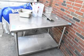 Stainless steel table with under shelf, containing various kitchen items. Used condition, located near brick wall.
