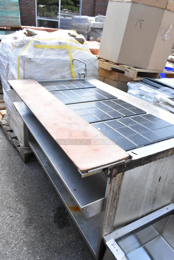Stainless steel commercial warming table with faucet, tiled surface, and metal shelving below; shows signs of wear.