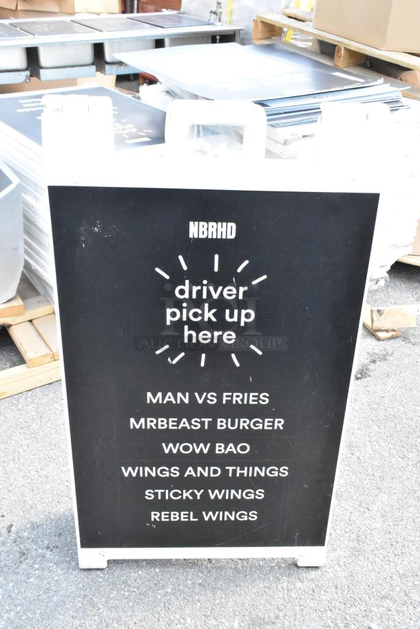 White A-frame sidewalk sign with "driver pick up here" text and food brands listed, slight wear visible on the surface.
