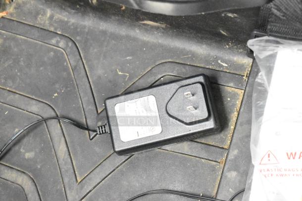 Close-up of a charger for the 2022 Pinghu 2000W Electric Children's Car, laying on a car mat, showing prongs and label.