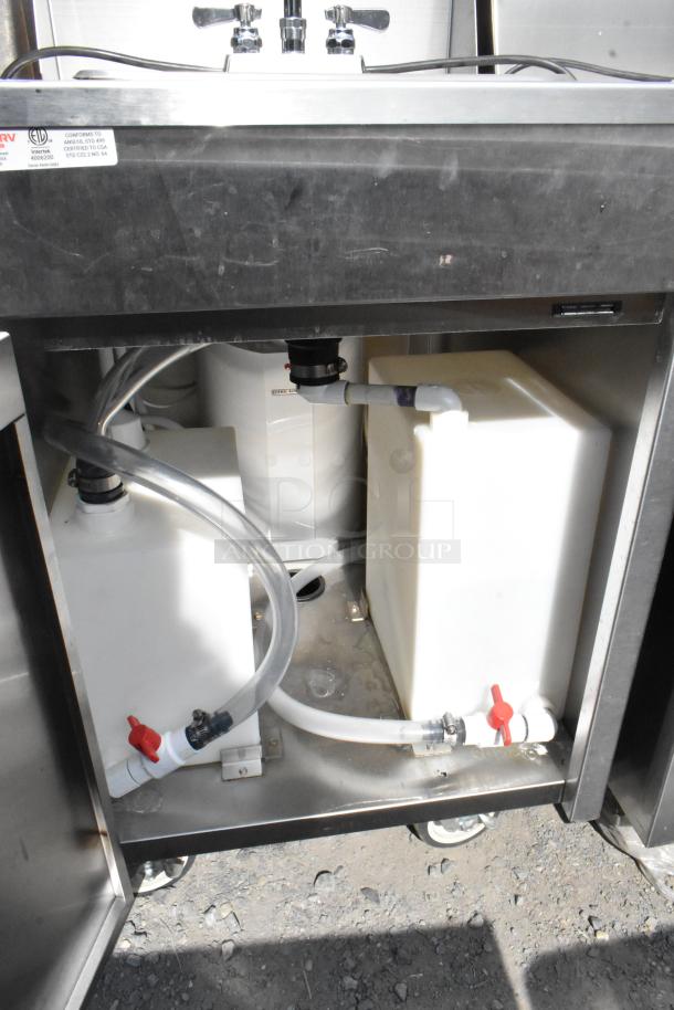 QualServ stainless steel portable sink with two faucets, visible plumbing, and commercial casters. Conforms to ANSI standards.