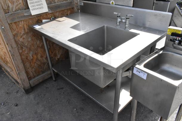 Stainless steel single bay sink with under shelf, dual faucets, and visible wear, ideal for commercial use.