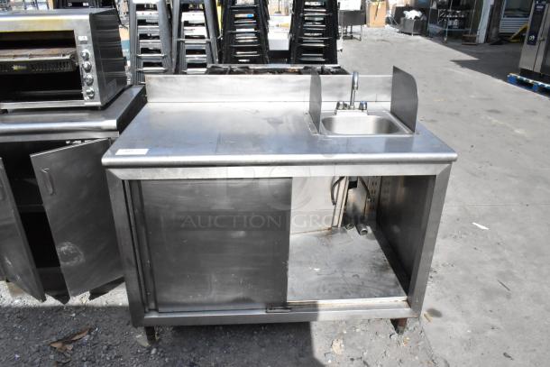 Stainless steel counter with sink basin, under shelf, and doors. Slight wear visible, ideal for commercial kitchen use.