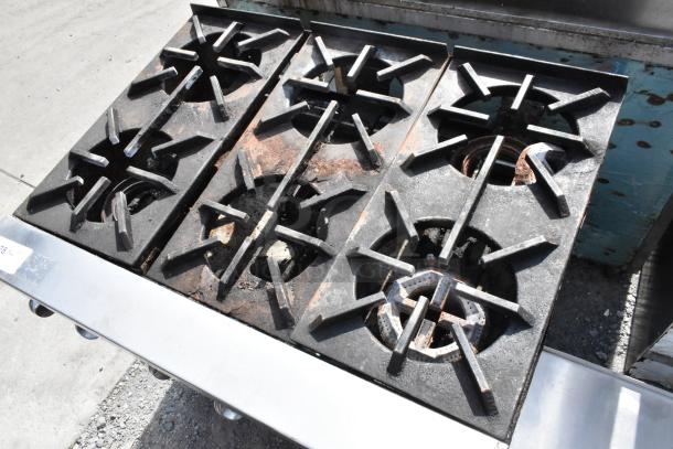 Stainless steel commercial gas-powered 6 burner range with under shelf, showing moderate wear. Ideal for professional kitchens.