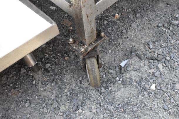 Stainless steel equipment stand with caster wheel, showing sturdy construction and light wear, on gravel surface.