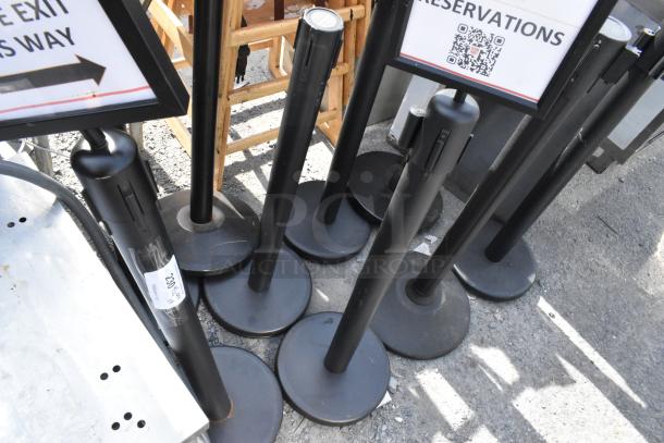 Nine black stanchions in used condition, grouped together, suitable for crowd control.