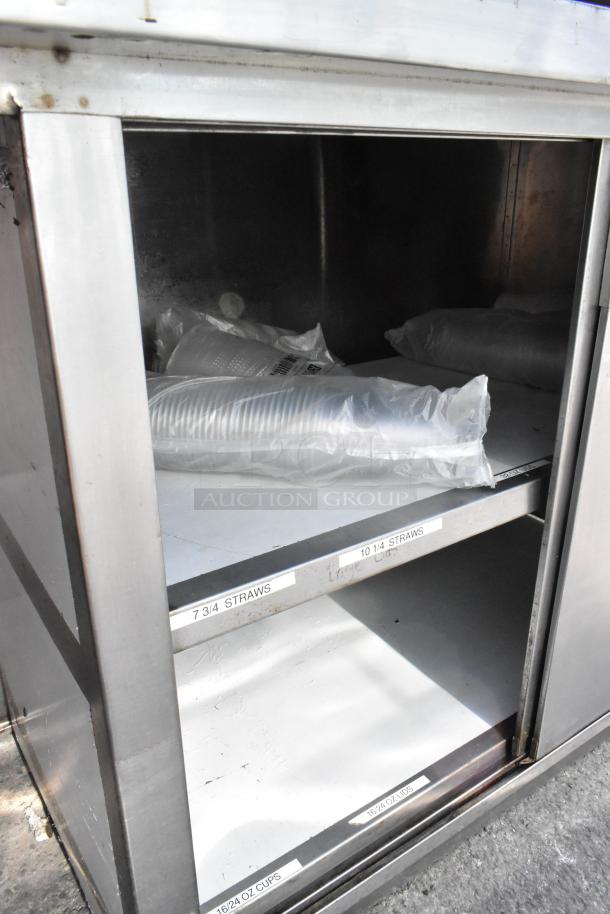 Stainless steel table with doors and under shelf, used, labeled for storing straws and cups. Surface shows wear.
