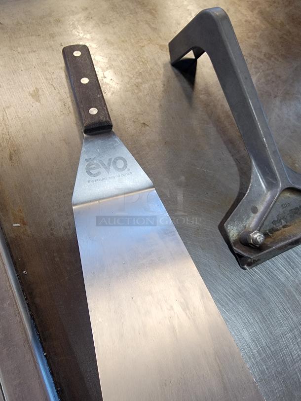 Evo spatula on stainless steel surface, showing "Evo, the natural way to cook" engraving, alongside metal press tool.