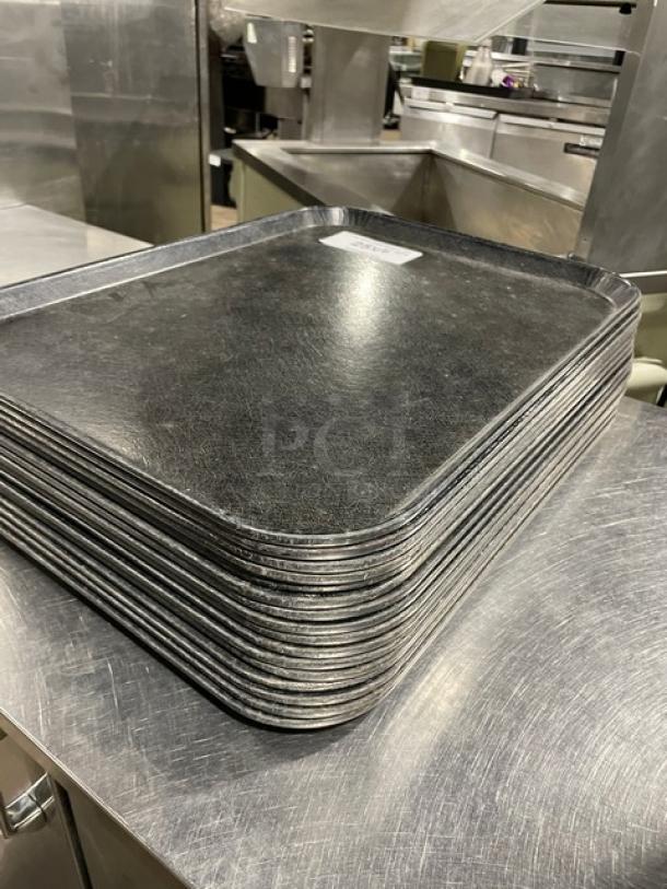 Stack of commercial black rectangular fast food serving trays on metal surface. Slight wear visible. Auction includes 16 trays.