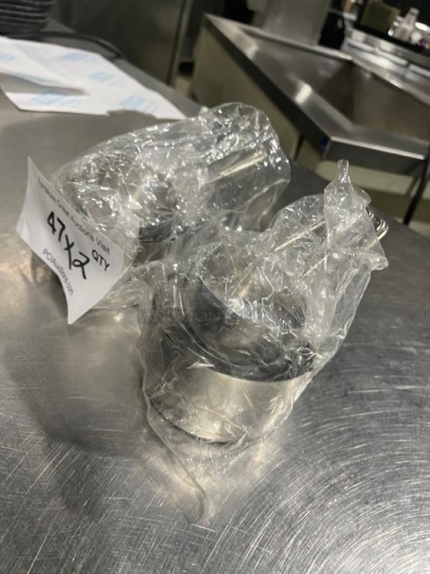 New commercial stainless steel measuring cup set wrapped in plastic. Includes tagging indicating "2X" for quantity.