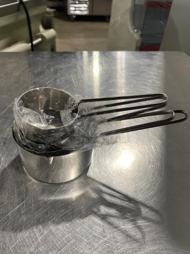 New commercial stainless steel measuring cup set, nested, wrapped in plastic, on metal surface.