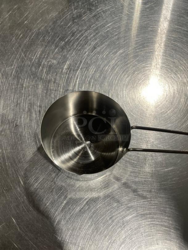 Stainless steel measuring cup on metal surface, new condition, part of commercial set, features marked measurements inside.