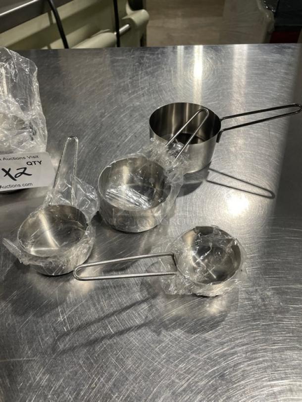 New commercial stainless steel measuring cup set, four pieces, wrapped in plastic, resting on a stainless table.
