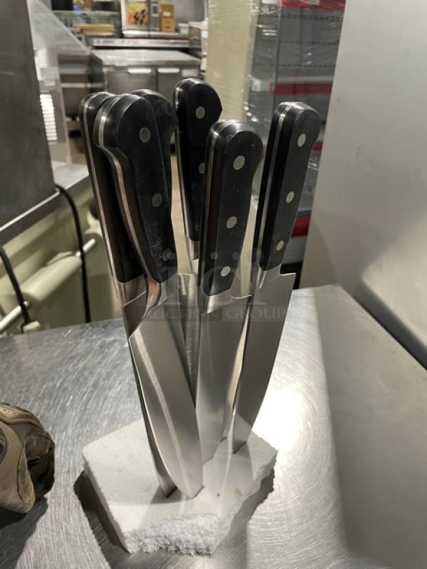 Set of new commercial stainless steel chef knives with black grip handles, displayed on a white block.