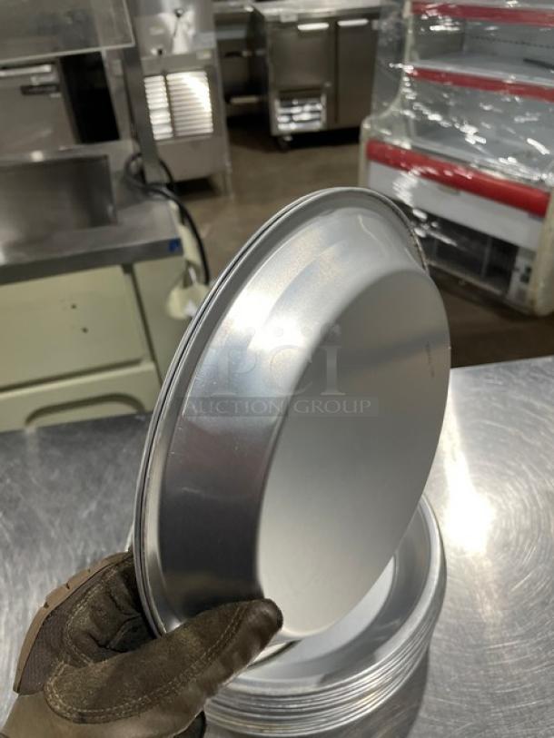 Stack of commercial round pie pans, metal, 19x quantity, used condition, held by gloved hand, on industrial kitchen counter.