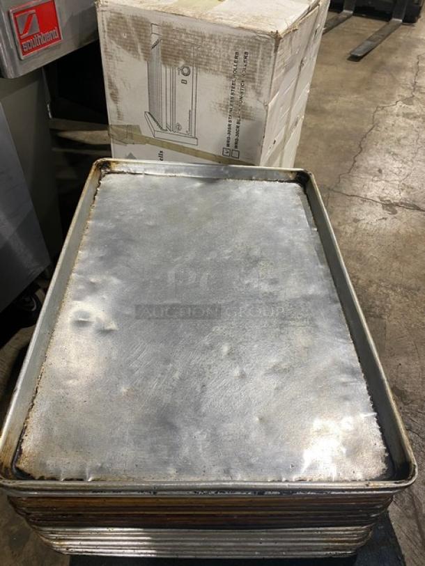 Stack of commercial full-size baking sheet pans, slightly worn condition, metal surface visible. Box in background.