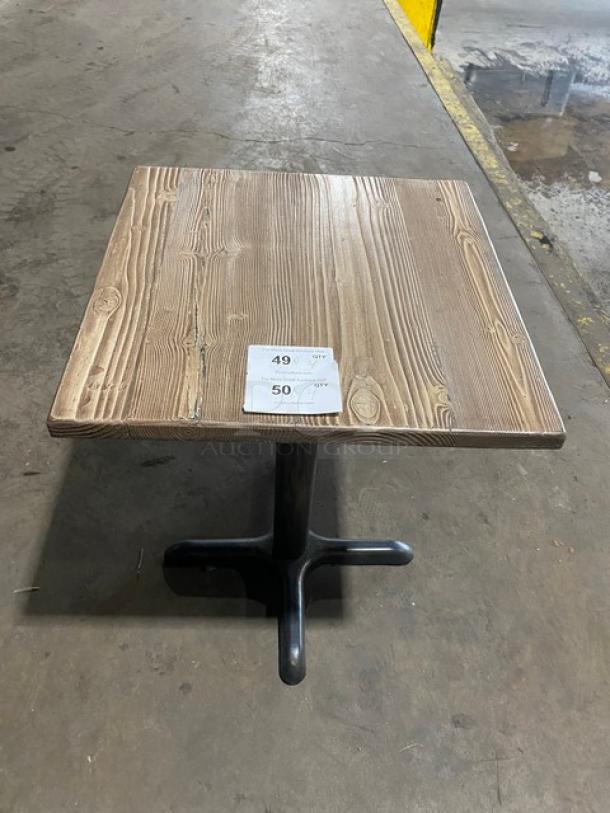 Commercial wooden square dining table with natural grain finish, black pedestal base, and auction tags visible on top.