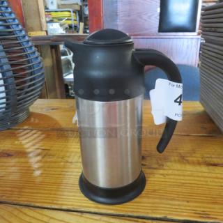 Stainless steel insulated creamer with black lid and handle on a wooden table, auction tag attached.