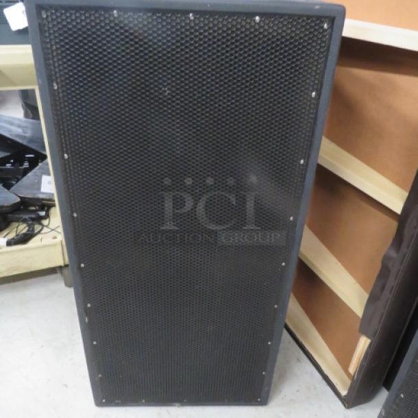 Large subwoofer speaker with black metal grille, rectangular frame, and visible screws; positioned upright on floor.
