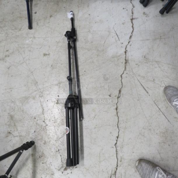 AKG mic stand, black, folded on a concrete floor. Features collapsible legs and adjustable height. Appears lightly used.