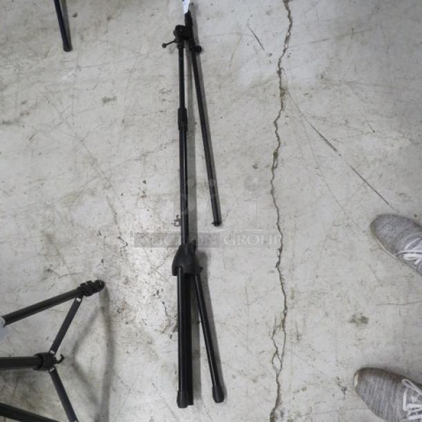 Adjustable black mic stand on a concrete floor, featuring a tripod base. Used condition with visible joints and adjustment knobs.