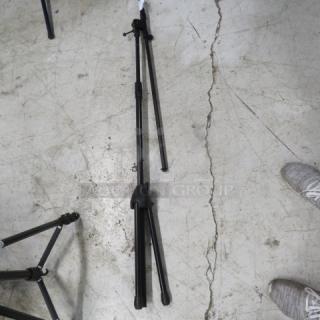 Adjustable black mic stand on a concrete floor, featuring a tripod base. Used condition with visible joints and adjustment knobs.